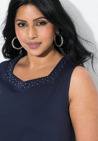 Marinblus utan ärmar med V-ringning som har metallstift längs halsringningen. Slät tyg med en lätt lyster.