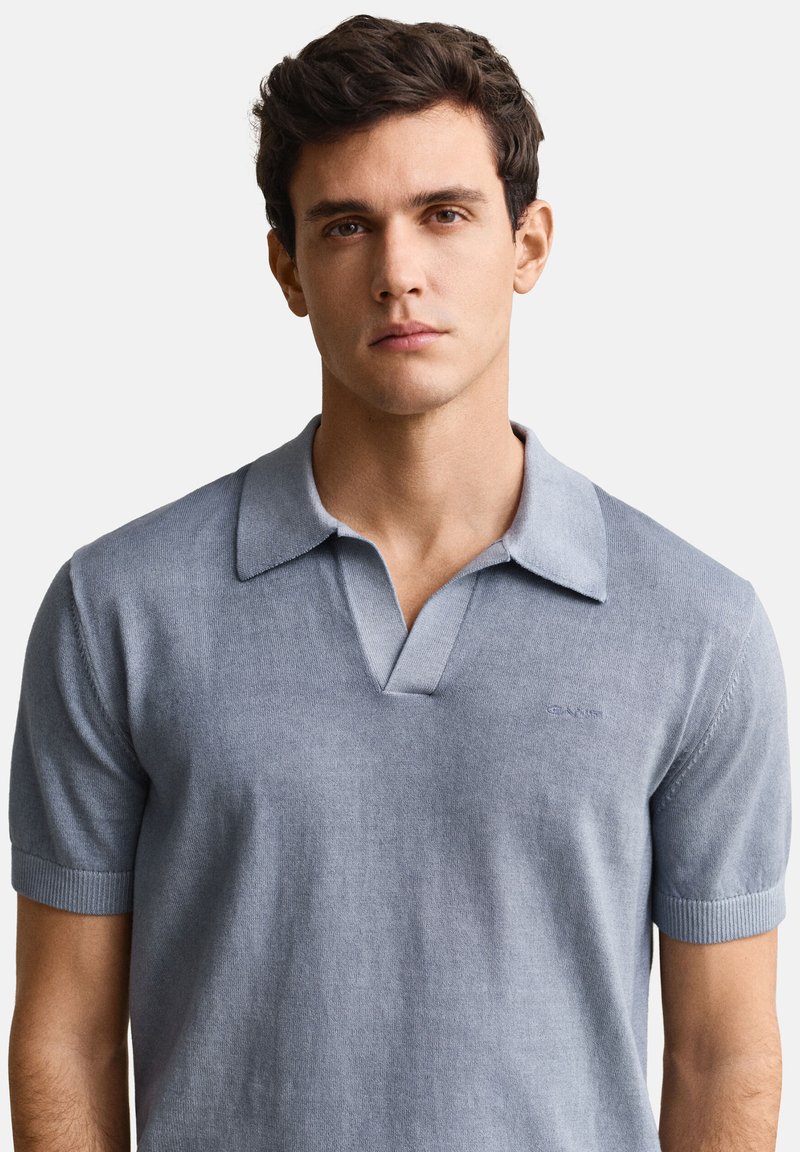 Joven con cabello oscuro y corto, vistiendo una camisa tipo polo de punto de manga corta color gris claro, mirando al frente sobre un fondo blanco liso.