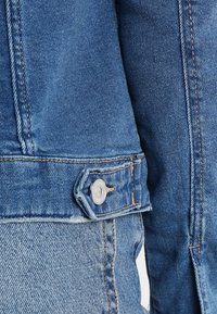 Gros plan d'une veste en denim bleue et d'un jean montrant des coutures cousues, un bouton en métal et la texture du tissu.