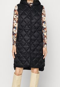 Černá prošívaná bezrukávová vesta s kapucí, vybavená předním zipem a šňůrkou. Nositá přes vzorovaný dlouhý rukáv a béžové boty.