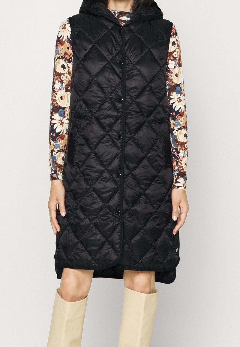 Černá prošívaná bezrukávová vesta s kapucí, vybavená předním zipem a šňůrkou. Nositá přes vzorovaný dlouhý rukáv a béžové boty.