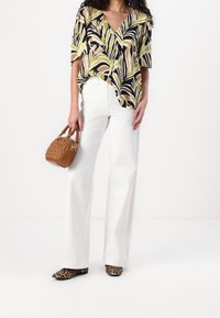 Maglietta a maniche corte e scollo a V con un motivo di foglie tropicali in verde, nero e beige, abbinata a pantaloni bianchi a gamba larga e a una piccola borsa marrone.