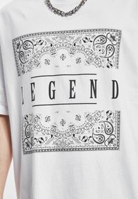 Hvit bomull t-skjorte med et svart paisley-mønster og ordet "LEGEND" i fet skrift over midten. Korte ermer og avslappet passform.