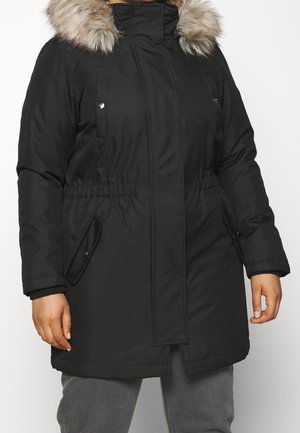 Persoon draagt een zwarte winterjas met een met bont gevoerde capuchon, voorzakken en elastische taille, gecombineerd met een grijze broek.