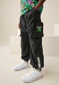 Fekete cargo nadrág állítható szárakkal, egy nagy oldalzsebbel, amelyen zöld Minecraft logó és fehér szöveg látható a lábszáron. Fehér sportcipővel viselve.