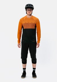 Langarm-Radsporttrikot in Orange und Schwarz, aus atmungsaktivem Material. Schwarze Shorts, Knieschützer und Radschuhe vervollständigen das Outfit.