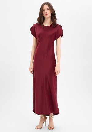 Femme debout portant une robe bordeaux en satin sans manches, longue jusqu'au sol, et des sandales dorées à talons hauts sur un fond blanc uni.