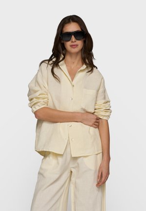 Kobieta w oversize’owej kremowej koszuli zapinanej na guziki i pasujących szerokich spodniach, z długimi brązowymi włosami i dużymi czarnymi okularami przeciwsłonecznymi.