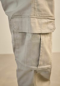 Pantalones cargo beiges con múltiples bolsillos, que cuentan con costuras reforzadas y una tela suave y texturizada. El diseño incluye puños doblados para ajustar la longitud.