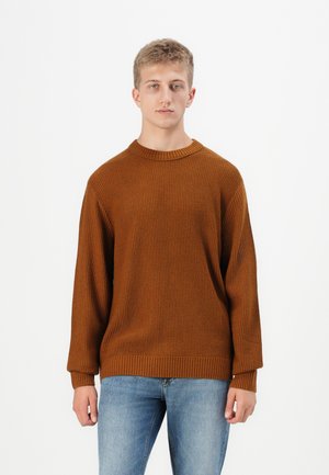 Pull en tricot marron avec un col rond, des épaules tombantes et des poignets côtelés. Associé à un jean bleu clair. Texture lisse, coupe décontractée.