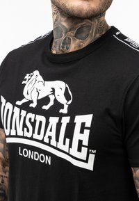 Černé bavlněné tričko s velkým bílým grafikou lva a textem "Lonsdale London" tučně. Design inspirovaný sportem se zvýrazněnými rameny.