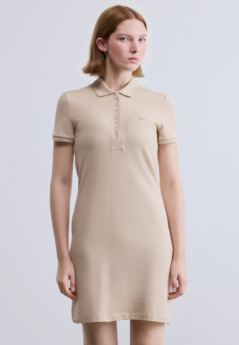 Μπεζ φόρεμα πόλο από απαλό ύφασμα, με κουμπιά στον γιακά, κοντά μανίκια και διακριτική λεπτομέρεια λογότυπου στο στήθος.