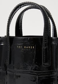 Musta patentoitu nahkalaukku, jossa on kaksi kahvaa, teksturoitu pinta ja kohokuvioitu kultainen logo, jossa lukee "Ted Baker London."