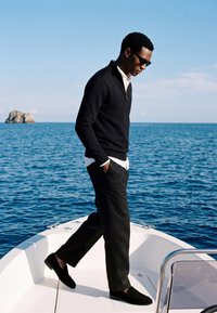 Homme portant un pull noir et un pantalon avec des lunettes de soleil, debout à la proue d’un bateau sur une mer bleue calme près d’une petite île rocheuse.