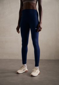 Marineblå leggings med høy midje har en hvit Nike-logo nederst på venstre ben; de er kombinert med beige joggesko.