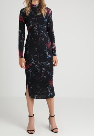 Donna che indossa un vestito midi a maniche lunghe nero con motivi floreali rossi e blu e sandali neri con tacco alto e cinturino alla caviglia.