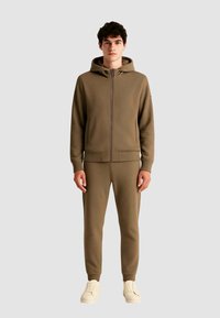 Set felpa e pantaloni khaki realizzato in morbido tessuto testurizzato. La felpa con cappuccio presenta una cerniera frontale, polsini a costine e un design minimalista.