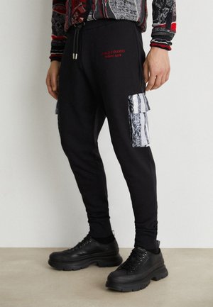 Osoba nosząca czarne joggery cargo z wzorzystymi bocznymi kieszeniami i czerwonym napisem "Carlo Colucci Milano 1978", w połączeniu z czarnymi masywnymi sneakersami.