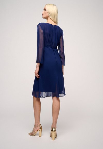Navy blue chiffon dress with long sheer sleeves, fitted waist, and flowy hemline. Back features gathered detailing and a keyhole closure.