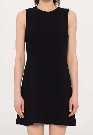 Femme portant une robe noire sans manches en ligne A, debout les bras détendus le long du corps sur un fond uni.