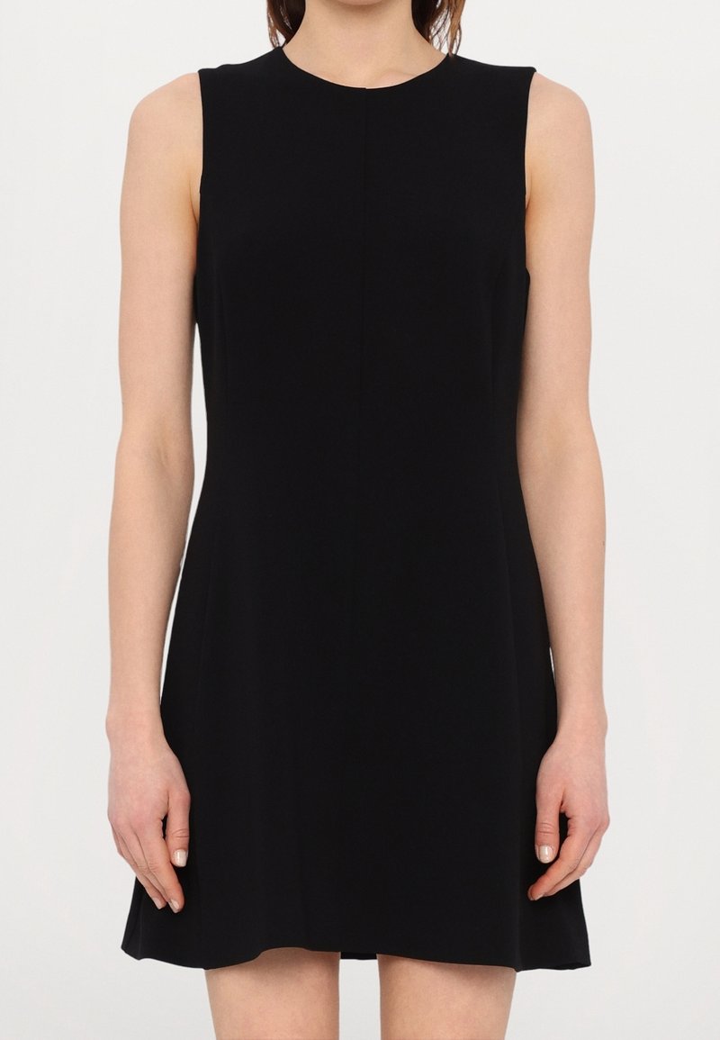 Femme portant une robe noire sans manches en ligne A, debout les bras détendus le long du corps sur un fond uni.