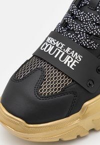 Zapatilla Versace Jeans Couture en negro y dorado con malla texturizada, cordones estampados y detalle de suela gruesa.