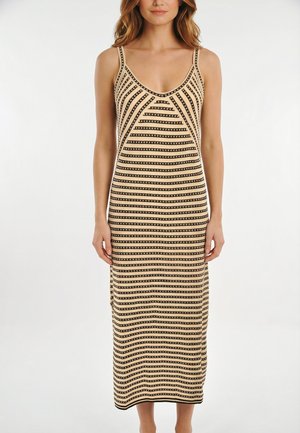 Vrouw in een mouwloze midi-jurk met beige en zwarte strepen en een ronde halslijn, staand tegen een effen witte achtergrond.