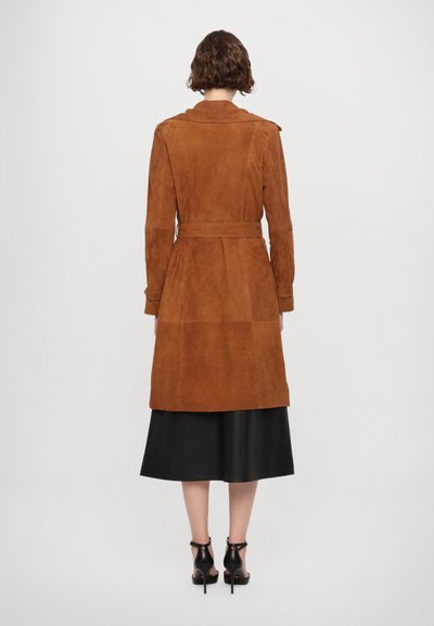 Cappotto trench in suede marrone con vita strizzata, maniche lunghe e uno spacco posteriore; abbinato a una gonna nera e tacchi. Texture liscia dappertutto.