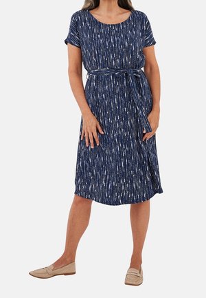 Mujer con vestido azul marino hasta la rodilla con rayas verticales blancas y cinturón de tela a juego, combinado con zapatos beige sin cordones.