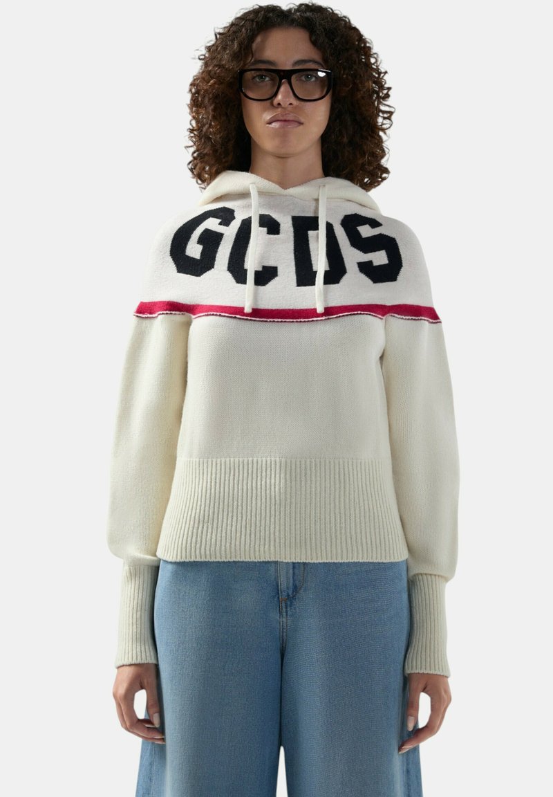 Donna con capelli ricci e occhiali che indossa una felpa bianca con la scritta "GCDS" e jeans blu a gamba larga su uno sfondo neutro.