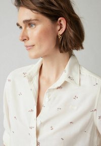 Camicia bianca con bottoni, decorata con piccoli motivi floreali nei colori tenui, caratterizzata da un colletto classico e una texture di tessuto morbida.