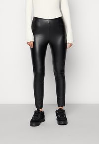 Schwarze kunstlederne Leggings mit einem eleganten, glänzenden Finish, ausgestattet mit einem hohen Bund und einem schmal zulaufenden Bein. Kombiniert mit einem weißen gerippten Top und schwarzen Sneakers.