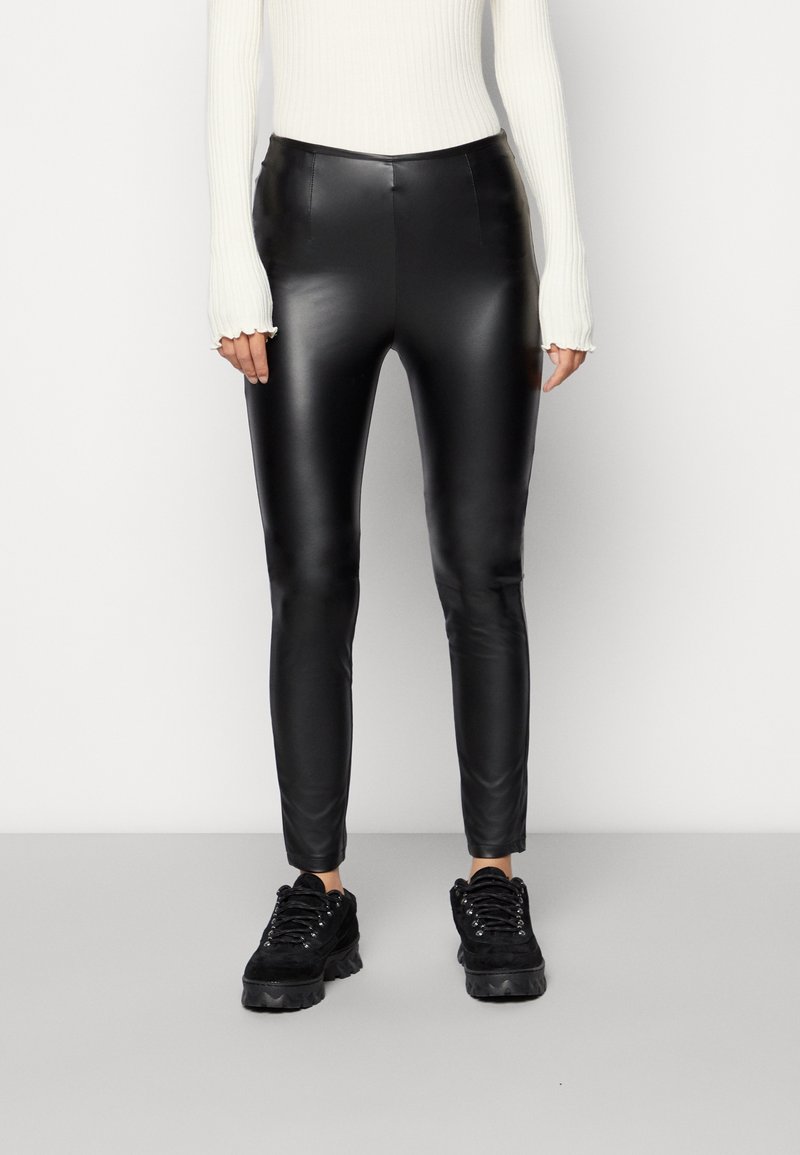 Schwarze kunstlederne Leggings mit einem eleganten, glänzenden Finish, ausgestattet mit einem hohen Bund und einem schmal zulaufenden Bein. Kombiniert mit einem weißen gerippten Top und schwarzen Sneakers.