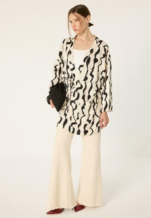 Mujer con chaqueta de estampado abstracto en blanco y negro, pantalones anchos color crema y zapatos rojos puntiagudos sosteniendo un bolso de mano negro.