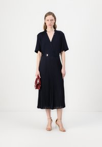 Navy pleated dress with a V-neckline, short sleeves, and a belted waist. Accessories include a small red handbag and nude high-heel sandals.