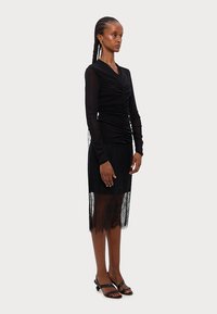 Black long-sleeve dress with a ruched bodice, featuring a lace hem and sheer sleeves, styled with black open-toe sandals.