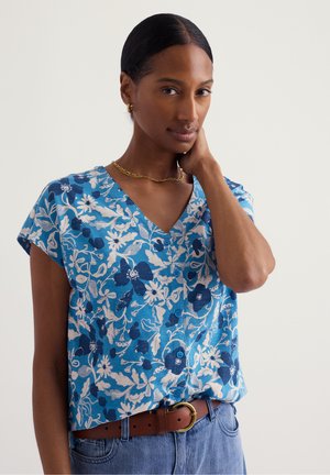 Žena s tmavými vlasmi, oblečená v modrej kvietkovanej košeli na gombíky, zlatom náhrdelníku a náušniciach, a modrých džínsoch s hnedým opaskom, ruka na krku.