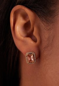 Gouden studoorbel met een rechthoekige, rozekleurige steen omringd door kleine heldere kristallen, gezet tegen een oor.