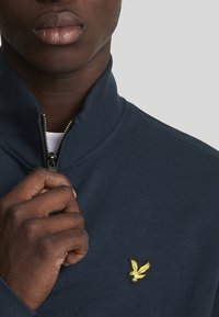 Felpa con zip blu navy, realizzata in tessuto morbido, dotata di colletto e di un piccolo logo di un'aquila gialla sul lato sinistro del petto.