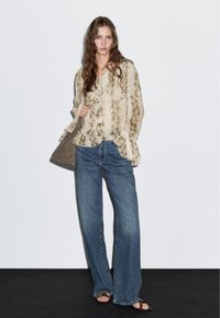Beige mönstrad blus med volanger, i kombination med högmidjade blå jeans. Modellen håller en taupe-färgad väska och bär bruna platåsandaler.