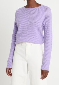 Pull en tricot texturé lilas avec un col rond et des manches longues côtelées. Présente un motif chevrons et une coupe décontractée. Associé à un pantalon blanc.