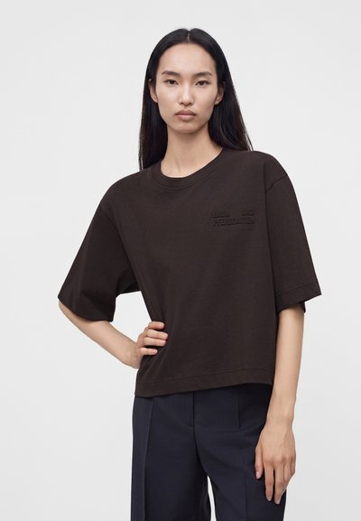 Femme aux longs cheveux noirs et lisses, portant une chemise ample à manches courtes marron foncé et un pantalon foncé, debout avec une main sur la hanche.