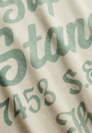 Bawełniana tkanina o kremowo-beżowym tle, z dużą zieloną typografią i misternymi literami. Tekst zawiera cyfry i stylizowane czcionki.