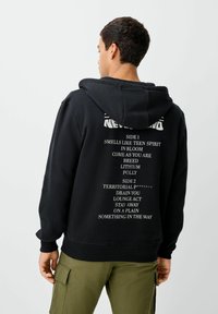 Vyras, vilkintis juodą gobtuv�ą su balta tekstu, nurodančiu dainų pavadinimus, stovi priešais paprastą šviesią foną, atsisukęs nuo kameros.