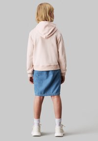 Felpa con cappuccio rosa chiaro e orlo a coste indossata sopra una gonna di jeans. Calzini bianchi e sneakers chiare completano l'outfit. Vista posteriore mostrata.