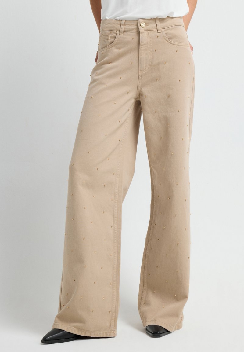 Beige wijde broek met kleine gouden kralenversieringen, gedragen met zwarte puntige schoenen en een witte top, getoond van taille tot voeten.