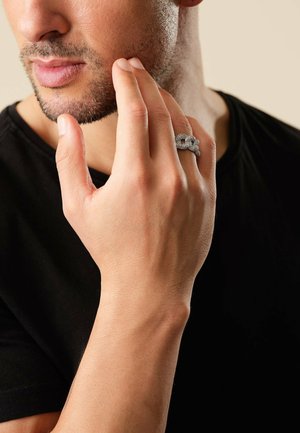 Silberner Ring mit strukturierten Details, getragen an der rechten Hand. Die Finger des Mannes berühren sanft sein Gesicht; er trägt ein schwarzes T-Shirt. Neutraler Hintergrund.
