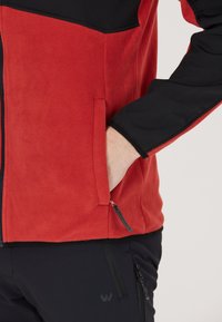 Röd och svart fleecejacka med dragkedja, sidofickor och kontrasterande paneler. Slät textur, strukturerad passform och hållbara design detaljer.
