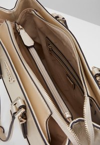 Beige nahkalaukku, jossa on kullanväriset yksityiskohdat ja musta somistus. Sisäpuolella on useita lokeroita, mukaan lukien vetoketjulliset taskut ja logotunniste.