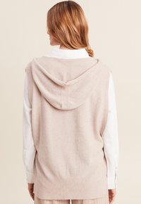Lichtroze mouwloze sweater vest met een capuchon, gemaakt van zacht materiaal; geribbelde details langs de randen; gelaagd over een wit overhemd met knopen.